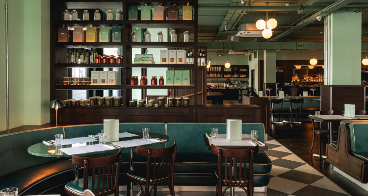Interiors of Lina Stores Manchester, featuring stocked deli shelves, teal velvet booth seating with contrasting pipe, marble tables and retro curved back chairs in dark wood.