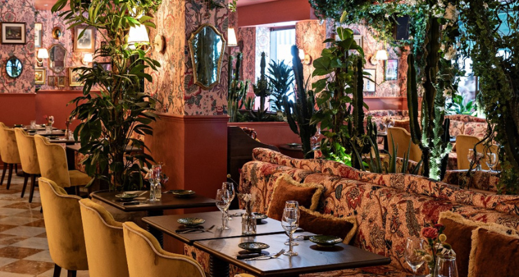 A picture of The Trafalgar's peach-coloured interiors, fronting giant tropical plants and mustard-toned velvet seats.