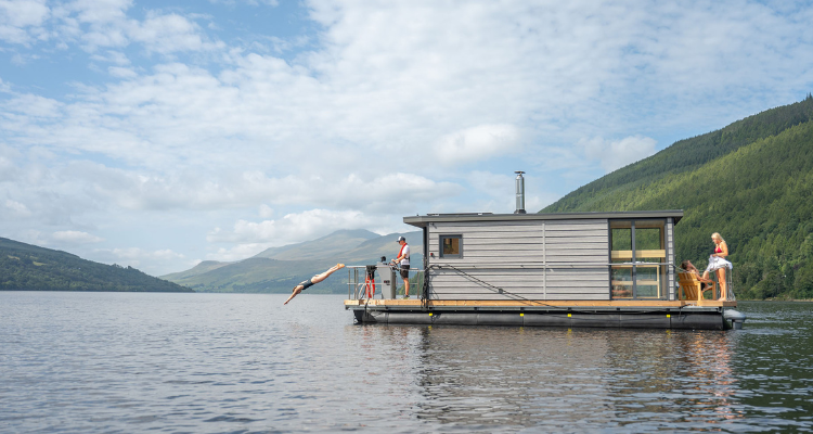 A person dives off a floating spa cruising Loch Tay, framed by Scottish Highlands.