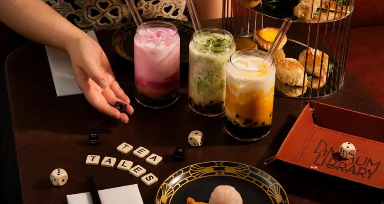 A hand with dice, three colourful, milky teas and dim sum in a afternoon tea birdcage.