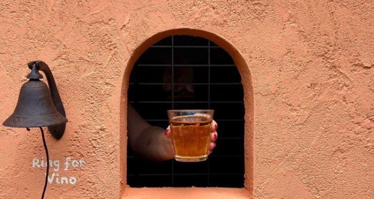 A hand passing a glass of wine through a hole in an orange wall. A bell is next to the hole with text that reads 'Ring for Vino.'