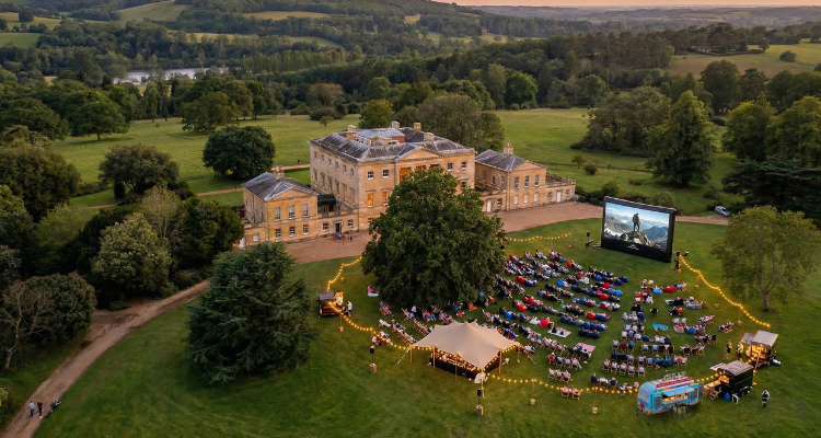 An open-air cinema set in a woodland with a historic house and rolling hills.