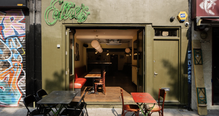 The exterior of a small bar with outdoor benches and a neon sign that says 'Ellie's'