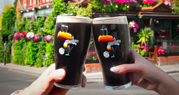 2 arms extended holding 2 pints of Guinness outside of a leafy pub.