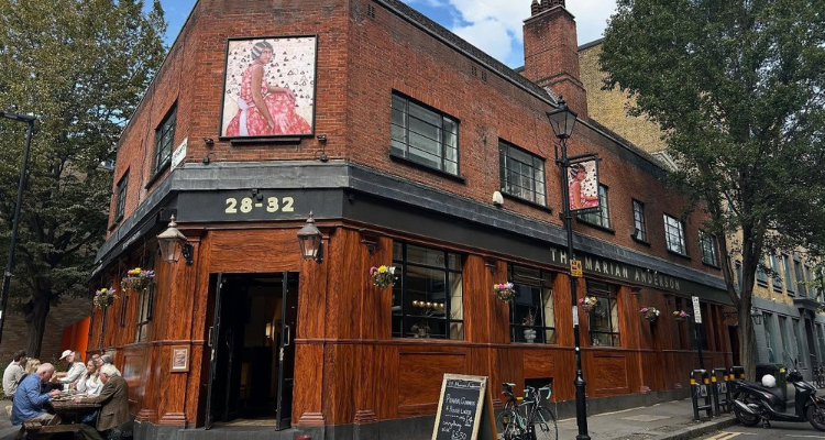 The exterior of a pub with red panelling, red brick, and people seated on a bench outside.