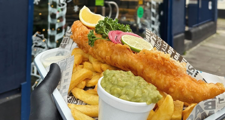 A gloved hand holding a takeaway container with fish and chips outside of a restaurant.