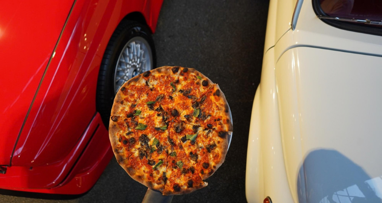 A pizza being held above two vintage cars. 