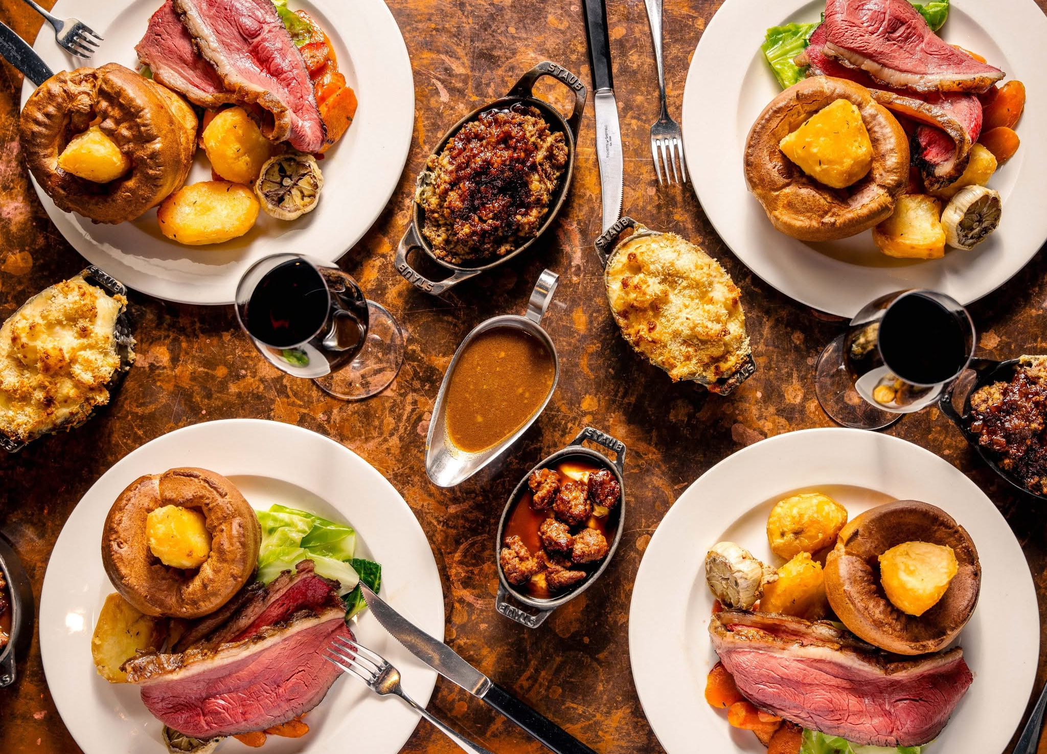 Four plates of Sunday roast, including rare beef, Yorkshire pudding, potatoes and green veg, with sides of cauliflower cheese and gravy.